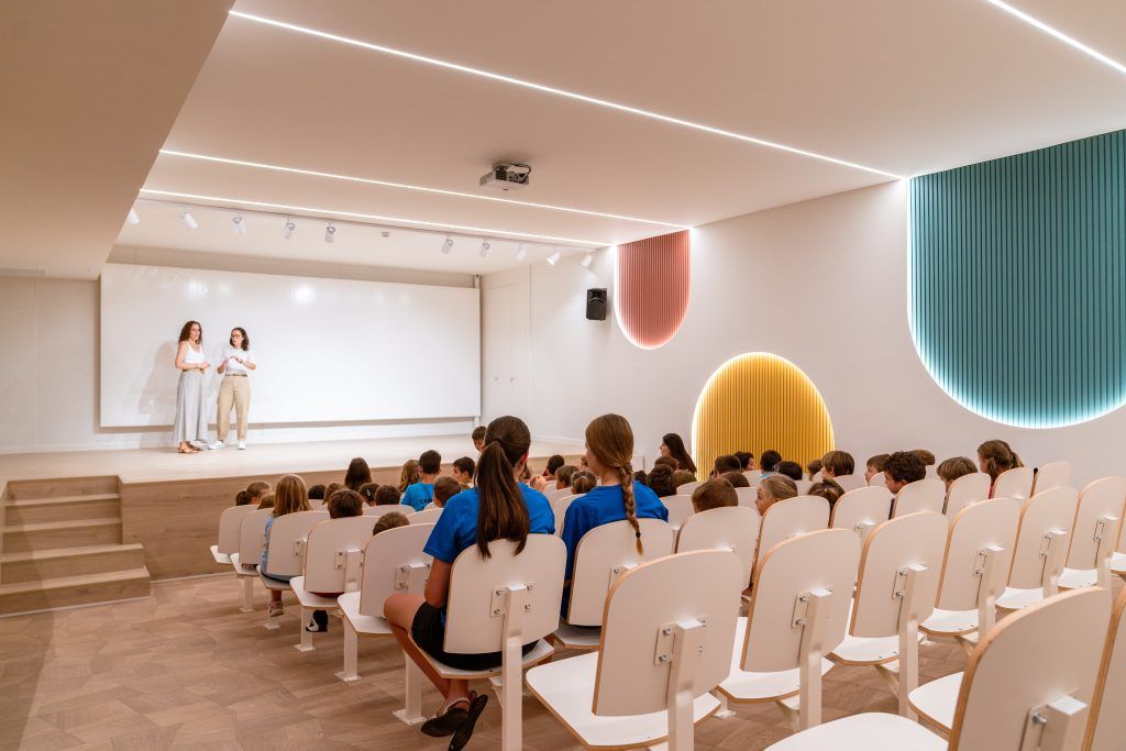 Auditorio escolar con estudiantes y escenario con iluminación moderna, ejemplo de iluminación en espacios educativos para presentaciones y aprendizaje.